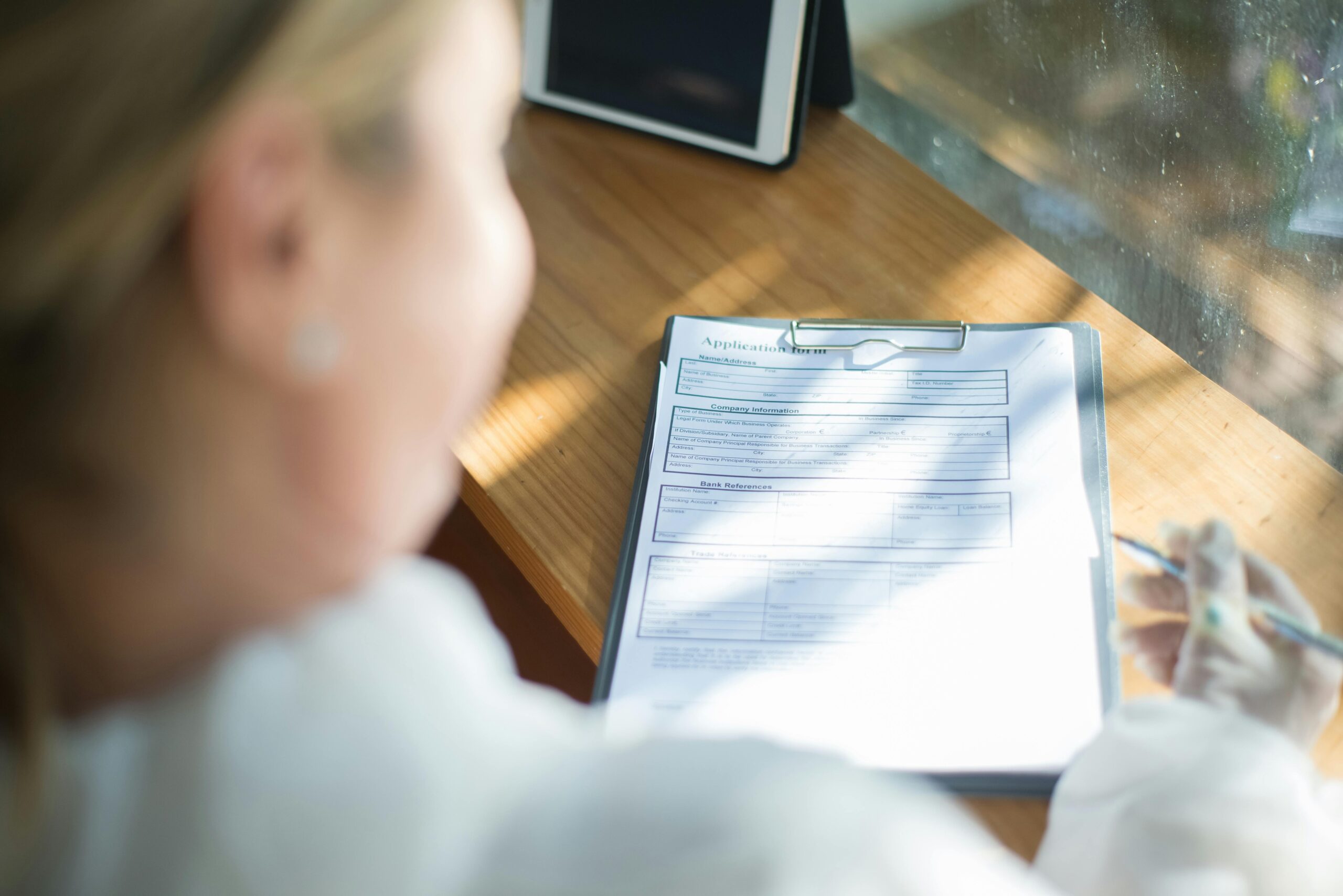A woman fills out an application form with a pen at a sunlit wooden desk.